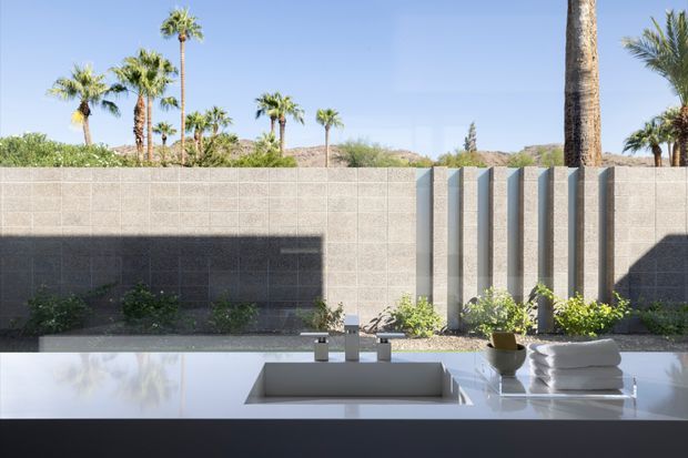 Minimalist bathroom vanity viewed from indoors, featuring a broad white countertop with an integrated rectangular basin and simple chrome fixtures. Beyond the glazing is a private landscaped courtyard with a textured concrete block wall, vertical projecting fins, desert planting, and palm trees. Strong sunlight creates crisp shadows across the wall and counter.