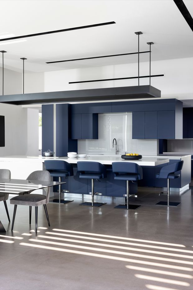 A bright, open-plan contemporary kitchen and dining area with deep navy-blue cabinetry, a large white island with four blue bar stools, integrated sink and cooktop, and a long suspended dark linear pendant light. The space has white walls and ceiling, polished grey concrete flooring, minimalist black track or linear lighting, and a dining table with grey upholstered chairs in the foreground. Strong sunlight creates linear shadows across the floor.