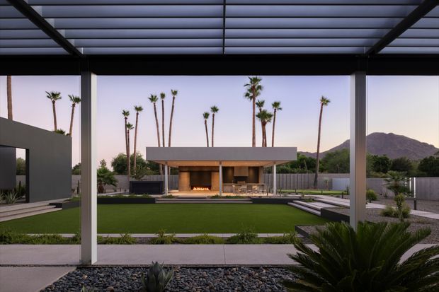 A refined outdoor living area viewed from beneath a large covered pergola with a translucent slatted roof and dark structural framing. The space opens onto a landscaped courtyard with a rectangular lawn, gravel beds, concrete paving, palms, and sculptural desert planting. A modern freestanding outdoor pavilion with a broad flat roof, timber-clad fireplace wall, and visible fire feature anchors the view. Mountains and tall palms form the distant backdrop.