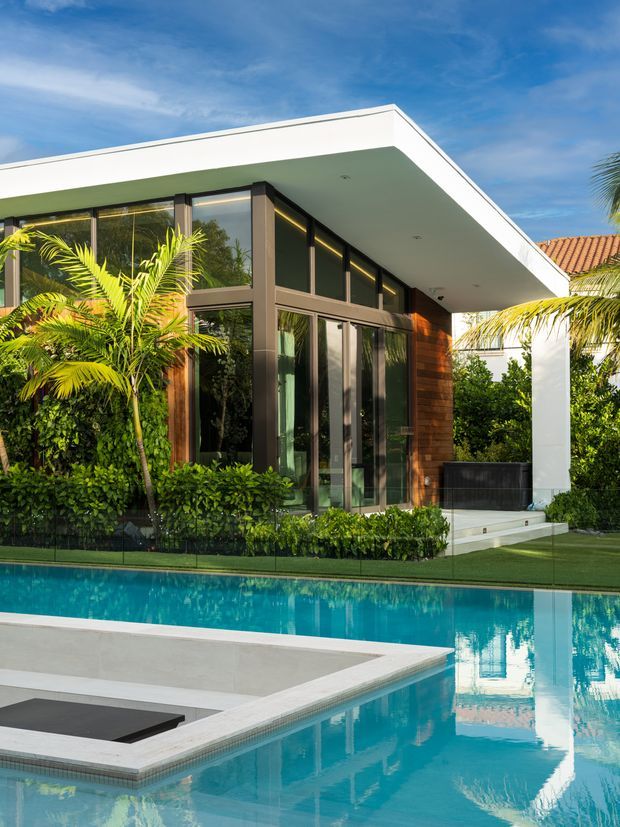 Exterior view of a contemporary flat-roofed pavilion beside a swimming pool, with expansive dark-framed glazing, warm timber wall cladding, white rendered soffits and lush tropical landscaping. The pool includes a pale paved edge and a sunken dark feature area, with strong reflections of the building in the water.