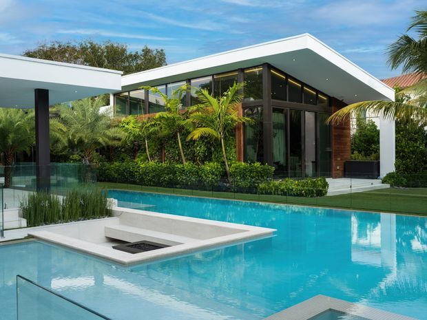A contemporary single-storey residence viewed from the swimming pool, featuring a sharply angled flat roof, extensive dark-framed floor-to-ceiling glazing, white rendered roof and wall planes, timber-clad sections, and lush tropical landscaping. The foreground contains a large turquoise pool with a raised white platform and built-in fire feature, plus a covered outdoor structure at left.