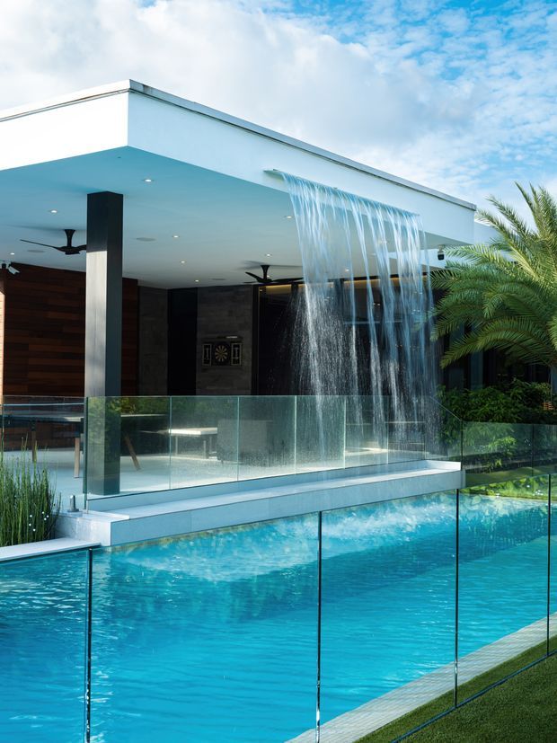 Contemporary poolside exterior featuring a large turquoise swimming pool enclosed by clear glass balustrades, a dramatic cascading water curtain, and a covered outdoor living area with white flat roof, recessed lighting, dark structural columns, timber wall cladding, ceiling fans, and tropical planting.
