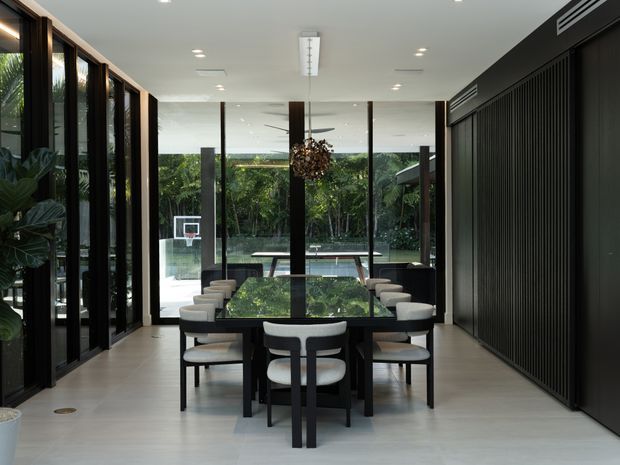 A refined contemporary dining room with a long dark dining table surrounded by upholstered chairs. Full-height glazed doors and windows open the room visually toward a landscaped garden and swimming pool, while a dark vertical slatted feature wall and dark-framed glazing create strong architectural contrast. A sculptural chandelier hangs above the table, and integrated ceiling lighting keeps the space bright and polished.