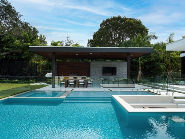 A contemporary outdoor pool area with a large turquoise swimming pool in the foreground, connected to a raised spa or shallow pool zone. Behind it is a covered open-air entertaining pavilion with a flat roof, dark timber ceiling, slender posts, outdoor dining furniture, a built-in television wall, and glass balustrades. Lush tropical landscaping surrounds the space.