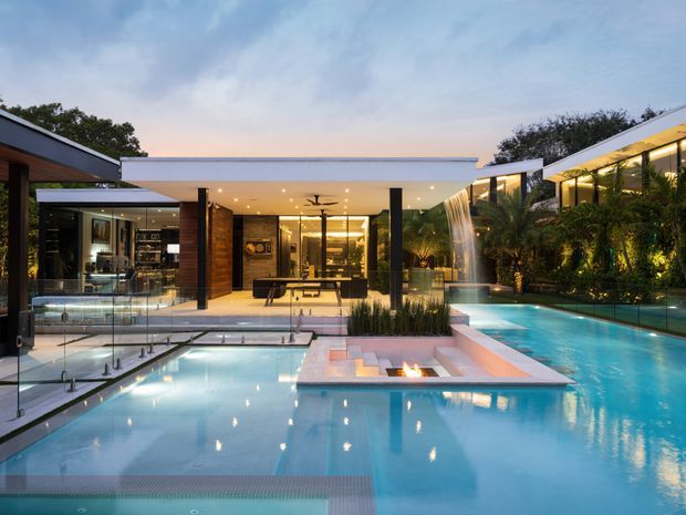 Contemporary luxury residence viewed from the pool area at dusk, with multiple connected flat-roofed wings, extensive floor-to-ceiling glazing, dark structural posts, warm timber-clad walls, and illuminated tropical landscaping. Several linked swimming pools and a raised central spa or fire feature dominate the foreground, while an open-plan living area is visible through the glazing.