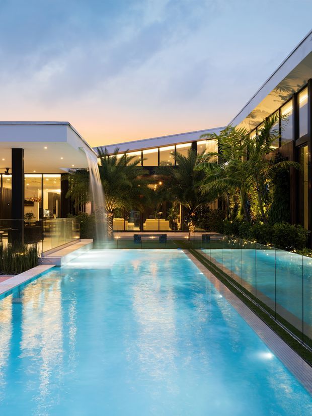 Twilight view of a contemporary courtyard residence centred around a long illuminated swimming pool. The pool is bordered by lush tropical planting, reflective glazing, dark-framed floor-to-ceiling windows, and crisp white flat-roofed pavilion forms. A narrow water feature or spillway flows into the pool near the left side, creating a resort-like atmosphere.