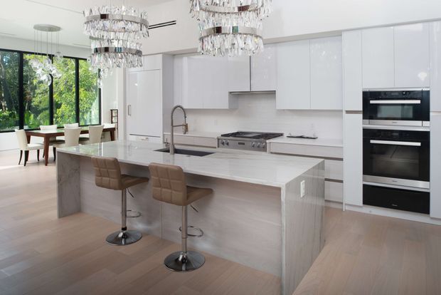 Bright, spacious contemporary kitchen with a large pale stone island and integrated seating for two. Gloss-white full-height cabinetry includes a concealed refrigerator and stacked ovens, with a stainless-steel range and sculptural curved faucet. A light neutral backsplash and pale timber flooring create a clean, refined palette. The kitchen opens to a dining area and leafy glazing, while ornate cascading crystal pendant lights provide a luxurious focal point.