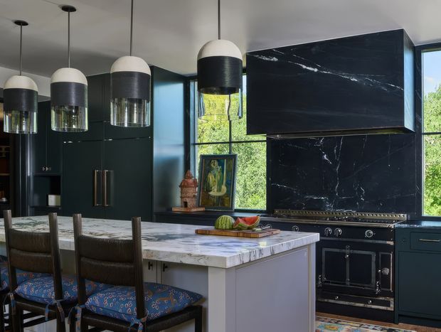 A sophisticated, moody kitchen with deep green-black cabinetry, a large marble-clad range hood and backsplash, and a professional-style black range. A pale marble island with waterfall edges and upholstered bar seating occupies the foreground, while oversized two-tone pendant lights hang above. Large windows bring in greenery and daylight, contrasting with the dark cabinetry and dramatic veined stone.