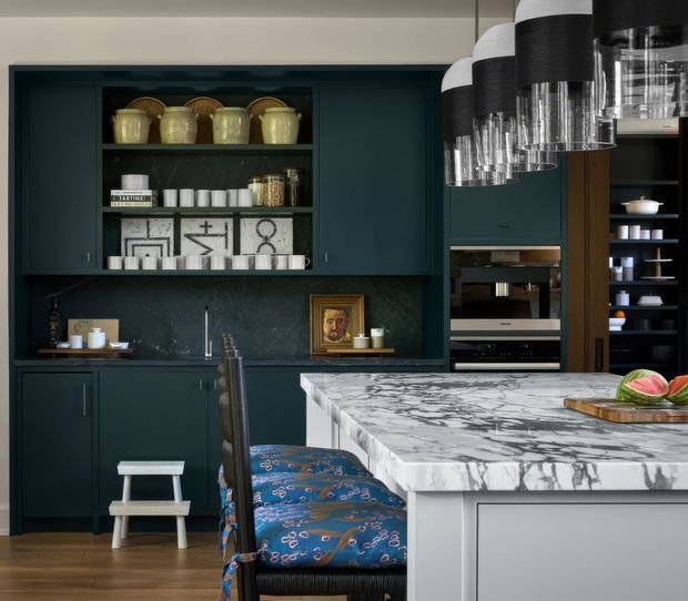 A sophisticated kitchen with dark teal built-in cabinetry, open display shelving, integrated appliances, a dark stone backsplash, and a large island topped with dramatic veined marble. Patterned upholstered island stools and sculptural pendant lights add decorative character, while warm timber flooring and brass-toned accessories soften the palette.