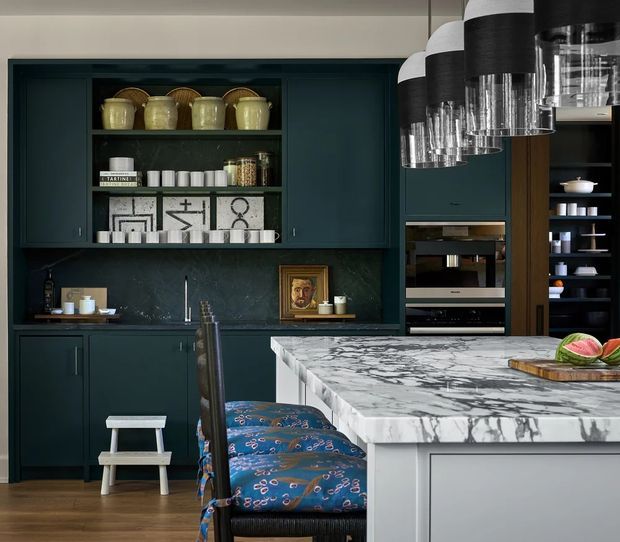 A refined, richly coloured kitchen with dark teal built-in cabinetry, open display shelving, integrated appliances, and a large white marble island with dramatic grey veining. Patterned blue upholstered bar stools sit at the island, while black pendant lights with clear glass lower sections hang above. Decorative ceramics, artwork, and tableware are arranged on the shelving and benchtop.