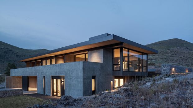 A contemporary two-storey concrete and glass residence set on a rugged, sloping hillside at dusk. The building has strong horizontal flat roof planes, extensive floor-to-ceiling glazing, exposed concrete walls, warm timber-toned interior lighting, and a partially recessed lower-level entry and garage-like opening. Surrounding hills and sparse rocky vegetation form the background.