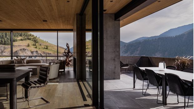 A contemporary mountain residence with floor-to-ceiling glazing opening from a refined living and dining interior onto a covered outdoor terrace. The space features a warm timber-lined ceiling, exposed concrete structural columns, dark metal-framed glazing, polished stone or concrete flooring, and understated modern furnishings. Sweeping mountain views dominate the background.