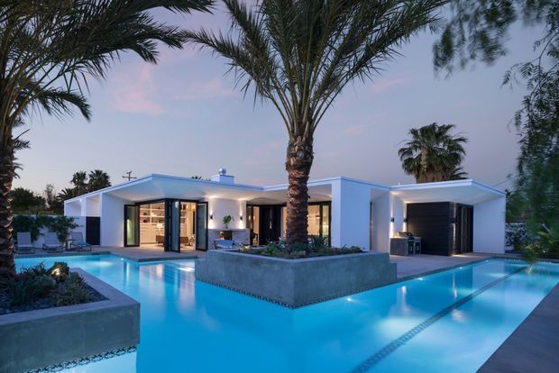 Twilight exterior view of a low-slung white modern residence arranged around a large illuminated swimming pool. The home features flat rooflines, extensive glazing, dark-framed sliding doors, a covered outdoor living area, integrated garage, concrete planters, and mature palm landscaping.