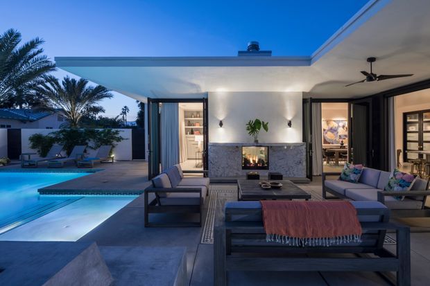 Evening view of a contemporary covered outdoor living area beside a swimming pool. The space features dark-framed open glazing, a built-in stone-clad fireplace wall, ceiling fan, lounge seating, coffee table, and patterned outdoor rug. Warm interior lighting is visible through the open doors, with landscaped palms and pool surrounds beyond.