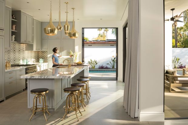 Bright, refined kitchen opening to an outdoor area with a pool visible beyond. The room features grey-green built-in cabinetry, a patterned geometric tile splashback, a large pale stone island with seating for four, brass pendant lights and stool bases, and full-height glazing or doors connecting to the exterior. A dining or lounge area is visible through the right-hand opening.