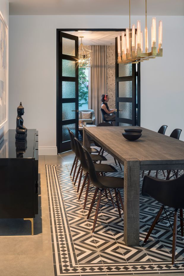 A refined dining room with a long rustic timber dining table surrounded by dark upholstered chairs. The room features a dramatic black-and-white geometric patterned floor runner or tiled floor design, black-framed double doors with textured glass opening to an adjoining sitting area, and a large sculptural brass-and-glass pendant chandelier. A dark sideboard with a Buddha sculpture sits along the left wall, while patterned wallpaper, drapery, and a desk area are visible beyond the doors.