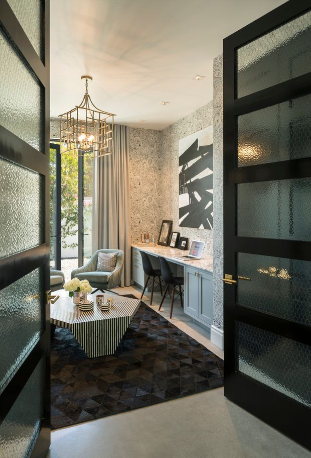 A refined, compact sitting room or study viewed through open black-framed doors with textured glass panels. The room features a patterned wallpapered wall, pale built-in desk cabinetry, two upholstered lounge chairs, a sculptural striped coffee table, a dark geometric rug, framed artwork, full-height curtains, and an ornate brass pendant light. Daylight enters through tall glazing, creating a sophisticated, layered interior.