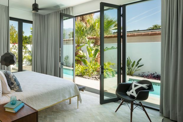 Bright contemporary bedroom with a low-profile upholstered bed, patterned accent cushion, timber bedside table, sculptural pendant light, and a black lounge chair. Full-height glazed doors open directly onto a private landscaped courtyard with a narrow turquoise pool, tropical planting, and rendered boundary walls. Floor-to-ceiling grey curtains frame the glazing, while pale geometric timber flooring adds texture.