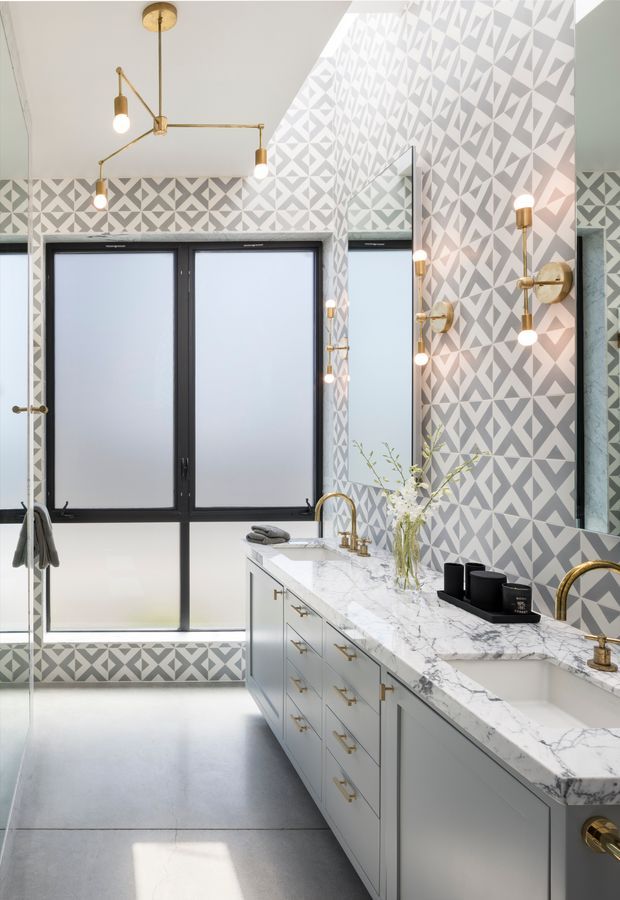 A bright, sophisticated bathroom featuring a long double vanity with pale cabinetry, white marble-look countertop, brass faucets and hardware, and black-framed frosted windows. Geometric grey-and-white patterned wall tiles wrap the room, complemented by brass globe wall sconces and a modern brass ceiling light. A large mirror reflects the vanity and patterned walls.