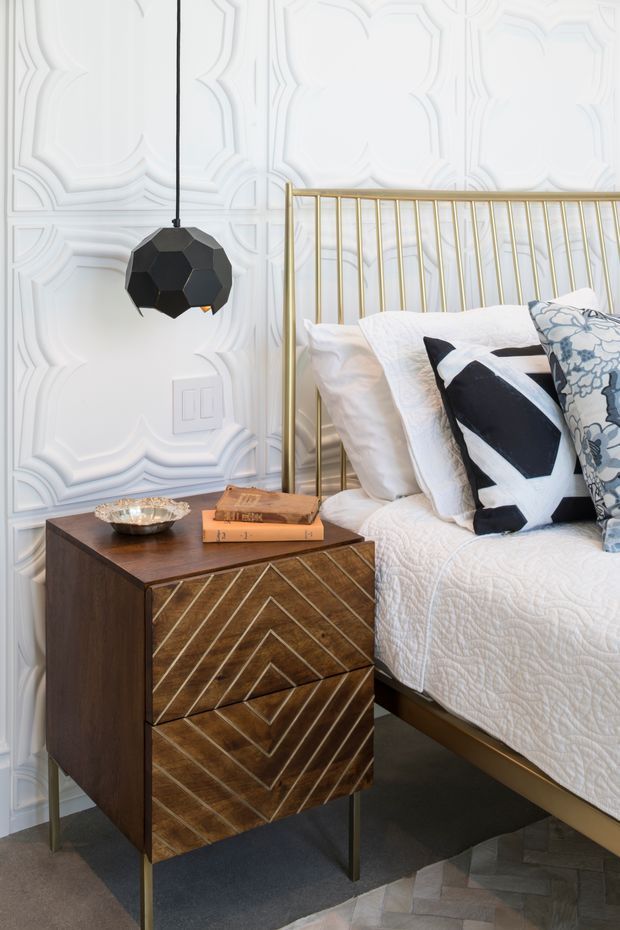 A close-up view of a stylish bedroom corner featuring a brass metal bed frame, white quilted bedding, patterned black-and-white and blue decorative pillows, and a dark timber bedside table with chevron-patterned drawer fronts and slim brass legs. The wall has ornate white raised geometric panel moulding, complemented by a black faceted pendant light and a small metallic bowl on the bedside table.