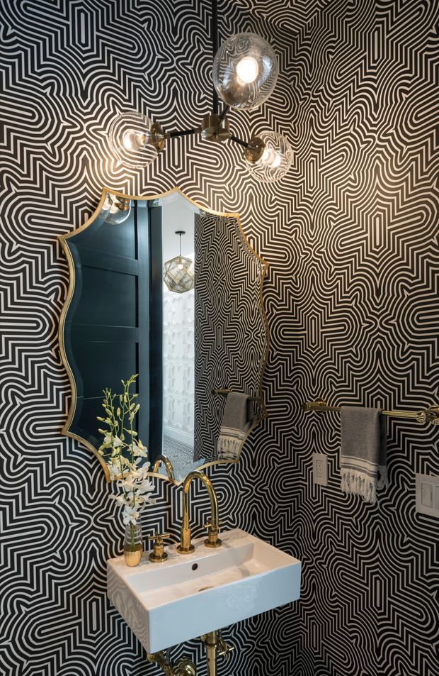 A compact, dramatically styled powder room with black-and-white geometric patterned wallcovering, a sculptural gold-framed wavy mirror, globe pendant lights, and a wall-mounted white basin with polished brass tapware. A dark blue reflective door or wall panel and patterned reflection add depth, while a small floral arrangement and folded towels provide limited decoration.
