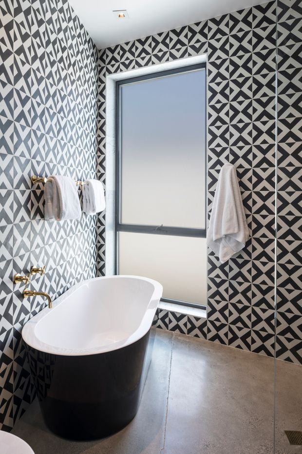 A compact, highly graphic bathroom featuring a freestanding black-and-white bathtub, dark metal-framed window, wall-mounted brass tapware, white towels, and bold geometric patterned wall tiles. The floor is a smooth grey concrete-like finish, creating a dramatic monochrome interior.
