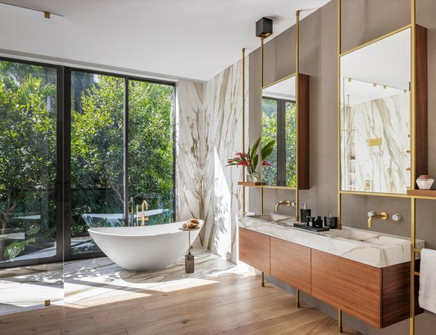 A bright, luxurious contemporary bathroom featuring a freestanding white bathtub beside floor-to-ceiling black-framed glazing, a large double vanity with warm timber cabinetry, pale stone benchtop, brass fixtures, and full-height veined marble-look wall surfaces. A large mirrored joinery wall reflects the tub and glazing, while lush greenery is visible outside.