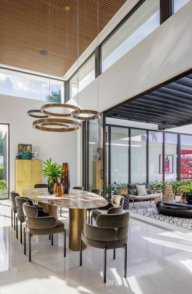 A bright, open-plan dining and living area with a double-height ceiling, full-height glazing, and a sophisticated contemporary interior. The dining zone features a large oval marble-look table with a metallic pedestal and upholstered dining chairs, beneath suspended circular ring lighting. A timber slatted ceiling, white walls, dark-framed glazing, indoor plants, and a nearby lounge with a sofa and coffee table complete the space.