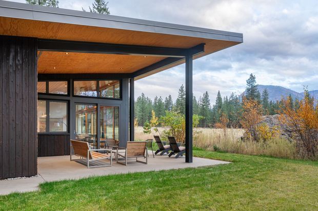 Exterior view of a contemporary single-storey pavilion-like home with a broad flat roof and deep covered patio. Dark vertical cladding contrasts with expansive black-framed glazing, while the soffit is lined in warm timber. Outdoor lounge furniture sits on a concrete terrace overlooking a grassy, wooded landscape with mountains in the distance.