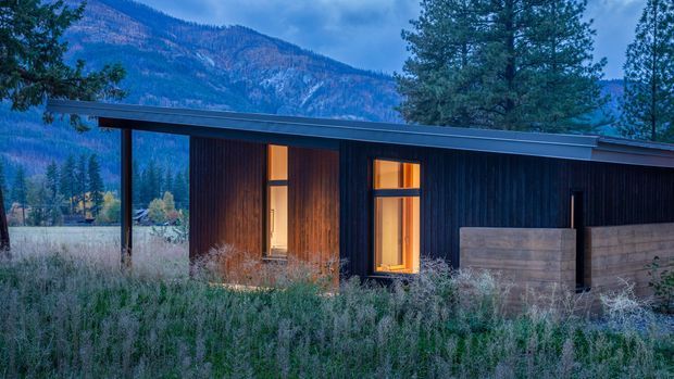 A low-profile contemporary single-storey dwelling set within a natural mountain landscape. The facade combines dark vertical timber cladding with warm timber entry detailing and large warm-lit windows, beneath a shallow low-pitched roof. A simple timber privacy fence and meadow planting sit in the foreground, with forested mountains beyond.