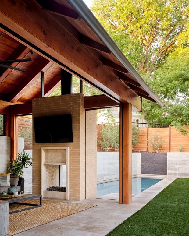A covered outdoor entertaining area beside a swimming pool, framed by a substantial timber gabled roof with exposed rafters and beams. The space includes a brick fireplace with a mounted television, outdoor seating, pale tiled paving, timber posts, and views to a landscaped courtyard and pool.