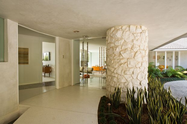 A bright, open-plan mid-century-inspired interior with a polished light floor, pale walls, full-height mirrored panels, and a prominent irregular stone-clad cylindrical column. The entry or circulation space opens toward a furnished living area, while a landscaped indoor planting bed and glazed courtyard are visible to the right. The space has a low, textured ceiling and strong indoor-outdoor connection.