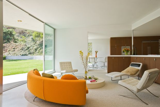 A bright open-plan mid-century-inspired living room with a curved orange sofa, cream lounge chairs, a round low coffee table, and a neutral carpeted floor. Floor-to-ceiling glazing opens to a green lawn and landscaped garden. Behind the living area is a compact dining zone and a dark timber kitchen with a central island, integrated appliances, and minimalist cabinetry. A large wall mirror expands the visual depth of the space.