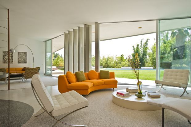 Bright, spacious mid-century-inspired living room with extensive floor-to-ceiling glazing opening toward a landscaped lawn and pool. The room features a low orange modular sofa, white tufted lounge chairs, a round white coffee table, sculptural floor lamp, neutral carpet, and a central row of vertical architectural fins dividing the living area from the adjacent seating space.