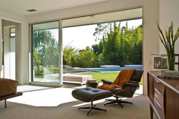 Bright mid-century-inspired living room with a classic lounge chair and ottoman, broad sliding glass doors, and a seamless connection to an outdoor pool and landscaped garden. The room has neutral carpet, white walls, expansive glazing, and a timber-toned low sideboard with indoor plants and artwork.