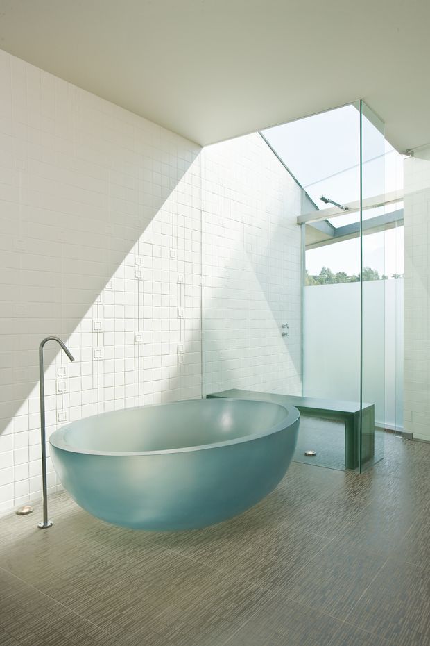 A bright, minimalist bathroom featuring a large freestanding oval bathtub, wall-mounted or floor-standing curved metal tap, white tiled walls, a glass-enclosed shower area, and a narrow integrated bench. The space has a skylight or clerestory glazing that creates strong geometric sunlight and shadow across the tiled walls. The floor is finished in small-format grey-brown mosaic tiles, with a soft pale blue-green bathtub as the main accent.