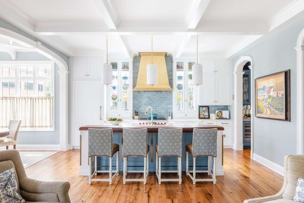 Bright, refined kitchen with a large central island and four upholstered stools, white custom cabinetry, a pale blue tiled backsplash, a distinctive gold-toned rangehood canopy, pendant lights, and exposed white ceiling beams. The open-plan space connects to a sitting area and includes traditional arched detailing, timber flooring, and abundant natural light.