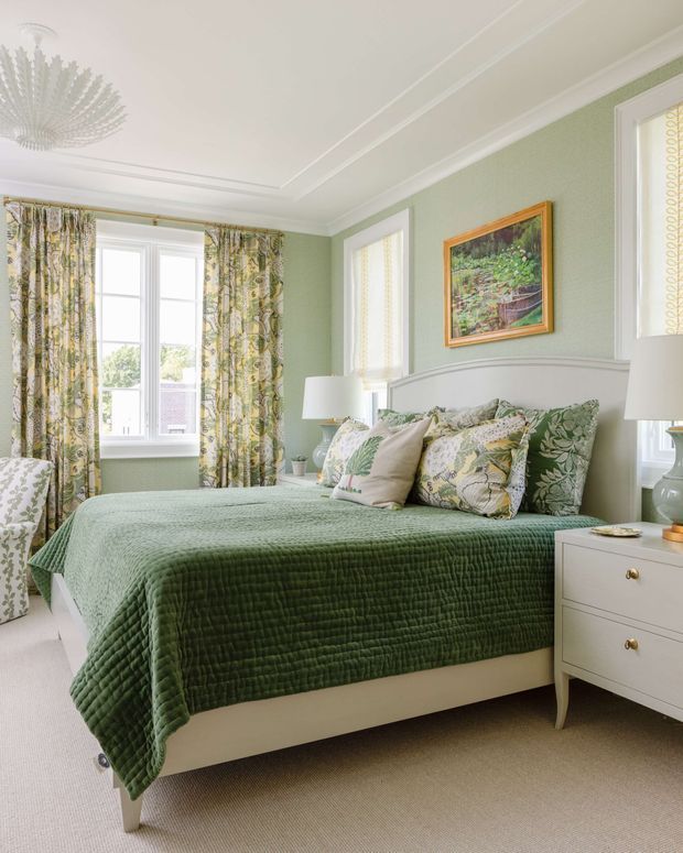 Bright, traditional-style bedroom with pale sage-green walls, white decorative ceiling moulding, a large upholstered bed with a curved headboard, layered green and botanical-patterned bedding, and matching floral curtains. White bedside furniture and lamps flank the bed, while a framed garden artwork and ceiling pendant add decorative detail. The room has generous windows and a calm, coordinated green-and-white palette.