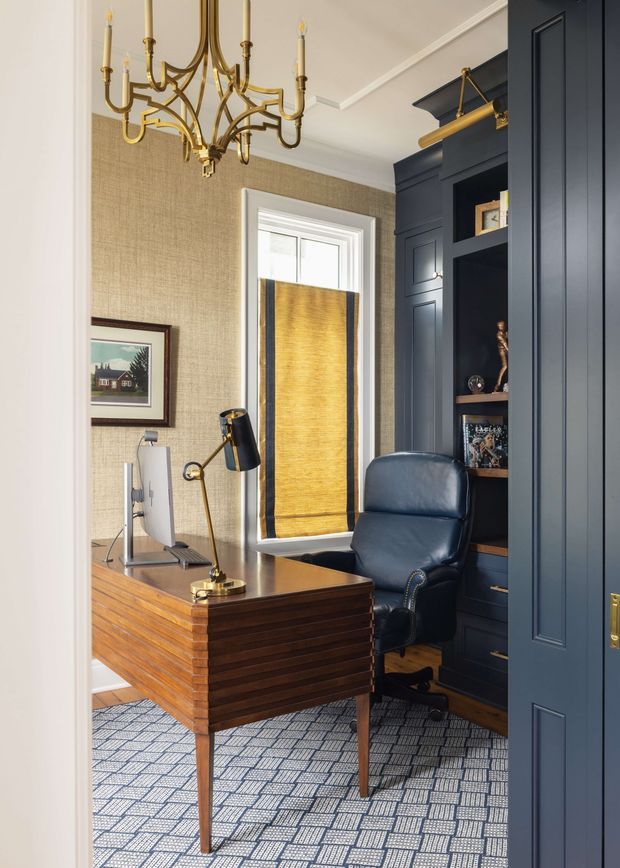 A refined, compact home office viewed through an open doorway. The room features a substantial dark timber desk with horizontal detailing, a navy leather office chair, dark blue built-in cabinetry with paneled doors and open shelving, woven neutral wallcovering, a patterned carpet, a brass articulated desk lamp, and an ornate brass chandelier. A window with a warm woven blind provides natural light.