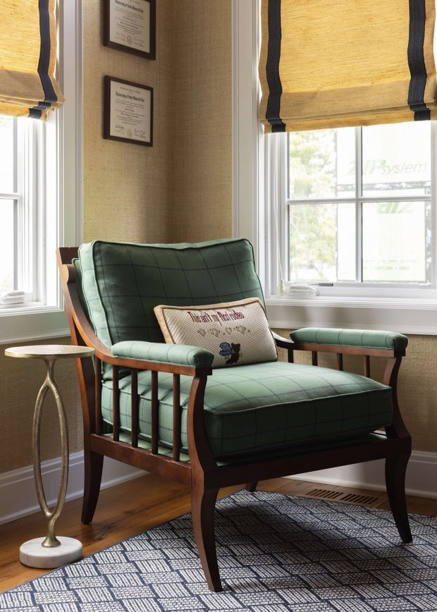 A refined sitting area centered on a deep green upholstered armchair with dark timber framing and curved arms. The chair is positioned between tall white-trimmed windows with woven bamboo or natural-fibre Roman blinds edged in dark trim. A slender metallic side table, patterned rug, timber floor, textured neutral wall covering, framed certificates, and a floor vent complete the room. The styling feels traditional with a tailored, heritage-inspired character and natural warm materials.
