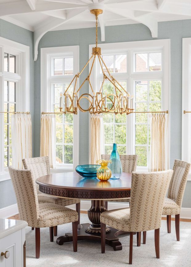 Bright, elegant dining room with pale blue-grey walls, white trim, decorative exposed ceiling beams, and large multi-pane windows. A round dark wood dining table is surrounded by upholstered dining chairs, with blue and amber glass vessels as centrepieces. A prominent ornate brass-gold chandelier hangs above the table, and light curtains frame the windows.