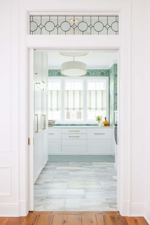 Bright view through a white-trimmed doorway into a compact, highly detailed kitchen or utility space. The room features white shaker-style cabinetry with brass hardware, a pale stone-look tiled floor, green patterned wallpaper and matching decorative countertop edging, three windows with striped Roman shades, and a ribbed circular pendant light. A decorative leaded-glass transom sits above the doorway, with warm timber flooring in the foreground.
