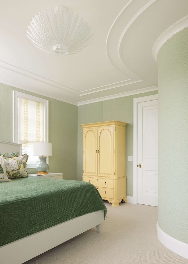 A softly lit bedroom with pale sage-green textured walls, a white bed dressed in a deep sage quilt, patterned cushions, and a yellow painted antique-style armoire. The room features a white paneled door, light carpet, detailed decorative ceiling mouldings, and an ornate white glass pendant light. A curved wall or column adds architectural interest on the right.