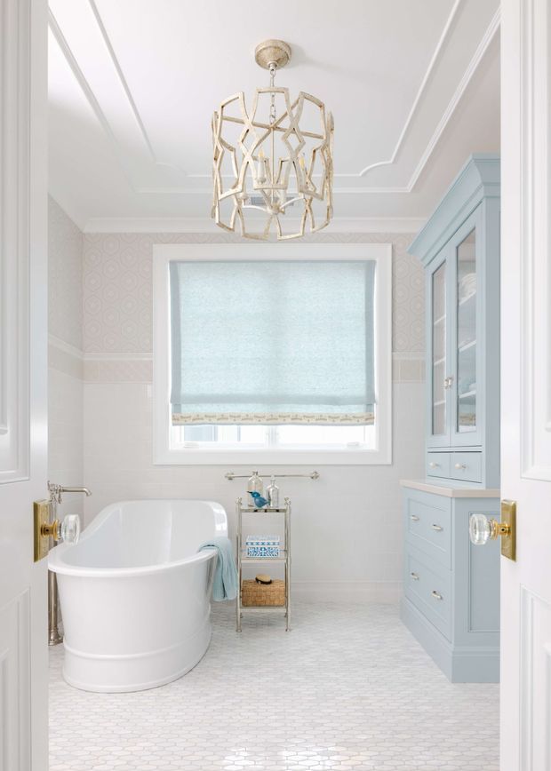 Bright, elegant bathroom with a freestanding white bathtub, pale blue built-in vanity and glass-front cabinetry, white patterned wall treatment, light hexagonal floor tiles, and a large window with a soft blue Roman blind. Decorative ceiling mouldings and an ornate gold-toned pendant light add a refined traditional character.