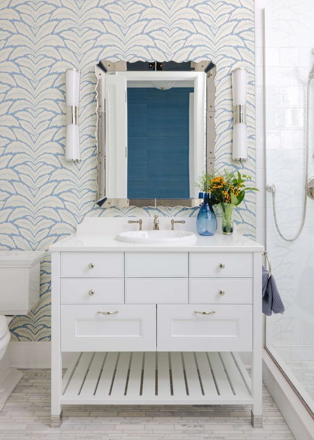 A bright, compact bathroom vanity area with a white freestanding shaker-style vanity, integrated white basin, chrome fixtures, and an open slatted lower shelf. The wall behind features blue and pale beige scallop-pattern wallpaper, flanked by vertical cylindrical wall sconces. A beveled mirrored cabinet hangs above the vanity, reflecting a deep blue-toned space. To the right is a white tiled shower enclosure with exposed plumbing and a handheld shower. The floor is finished in small light rectangular tiles, and blue glass vessels with flowers add colour.