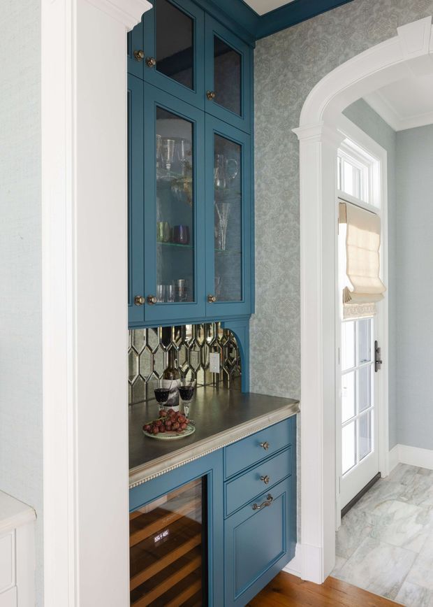 A refined built-in beverage bar and glassware cabinet set within a bright transitional interior. Deep teal-blue cabinetry with glass-front doors, brass-toned hardware, a reflective metallic geometric backsplash, stone benchtop, and integrated wine storage sit beside a white-trimmed arched opening and glazed exterior door. Pale patterned wallpaper and marble-look flooring add classic decorative detail.
