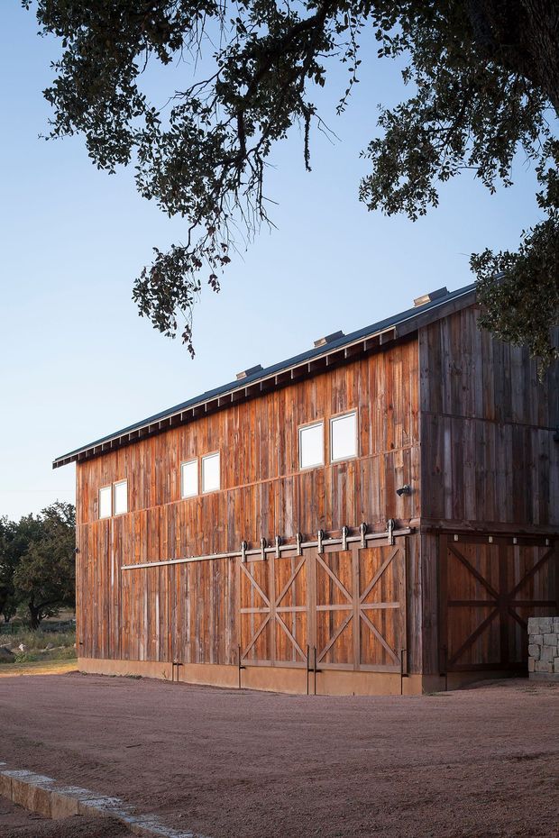 Exterior view of a large rustic timber barn or agricultural outbuilding with weathered vertical wood cladding, oversized timber barn doors, small rectangular windows, and a simple pitched roof. The building sits in an open gravel forecourt beneath mature trees.