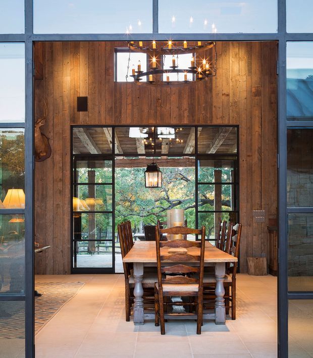 A rustic dining room framed by extensive dark metal glazing, featuring a large solid timber dining table with carved pedestal legs and matching wooden chairs. The room has warm vertical timber wall panelling, a large internal steel-framed glazed opening with views to a leafy outdoor area, exposed timber ceiling elements, and multiple suspended pendant and chandelier-style lights. A tiled floor and adjacent glazed spaces reinforce the open, industrial-rustic character.