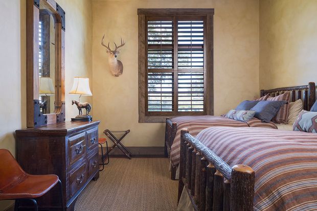 Rustic bedroom with twin beds featuring dark log-style timber frames and striped bedding. The room has warm textured plaster walls, a dark timber dresser with mirror, plantation shutters, patterned carpet, leather seating, and rustic deer decor, creating a cozy lodge-inspired atmosphere.
