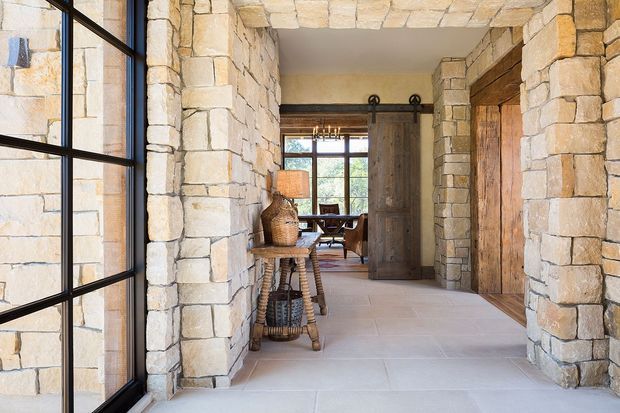 A rustic stone-lined interior entry hall with large black-framed glazing on the left, pale stone masonry walls and columns, a large pale tiled floor, and substantial timber doors. A sliding barn-style door opens toward a room beyond with a dining table and chairs. The space combines rugged natural stone, warm timber, and dark metal detailing.